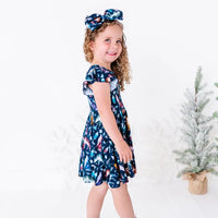 Side view of a child smiling over the shoulder in a flutter sleeve twirl dress with a matching bow headband, in a navy print of geometric wolves, triangle pine trees, and snowflakes.