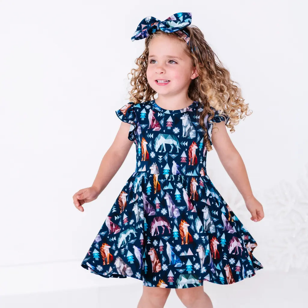 A child dancing with arms stretched out and the skirt swinging, in a flutter sleeve dress in a navy print of geometric wolves, triangle pine trees, and snowflakes.