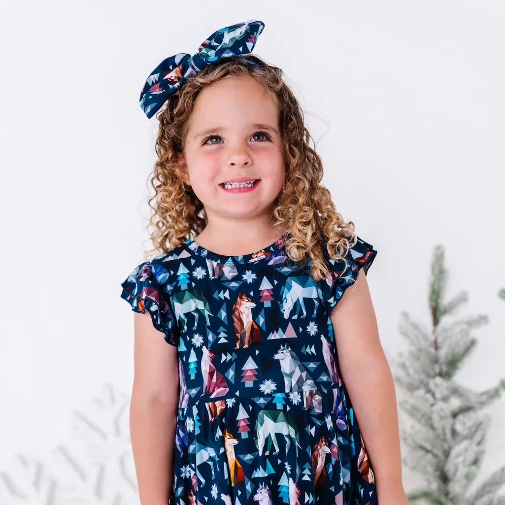 A child with curly hair smiling at the camera in a flutter sleeve twirl dress and matching bow in a navy print of geometric wolves, triangle pine trees, and snowflakes.