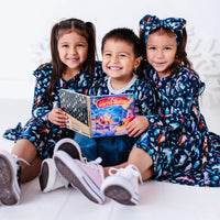 Three siblings sitting with a storybook and smiling, in two long sleeve twirl dresses and a pocket tee in a navy print of geometric wolves, triangle pine trees, and snowflakes.