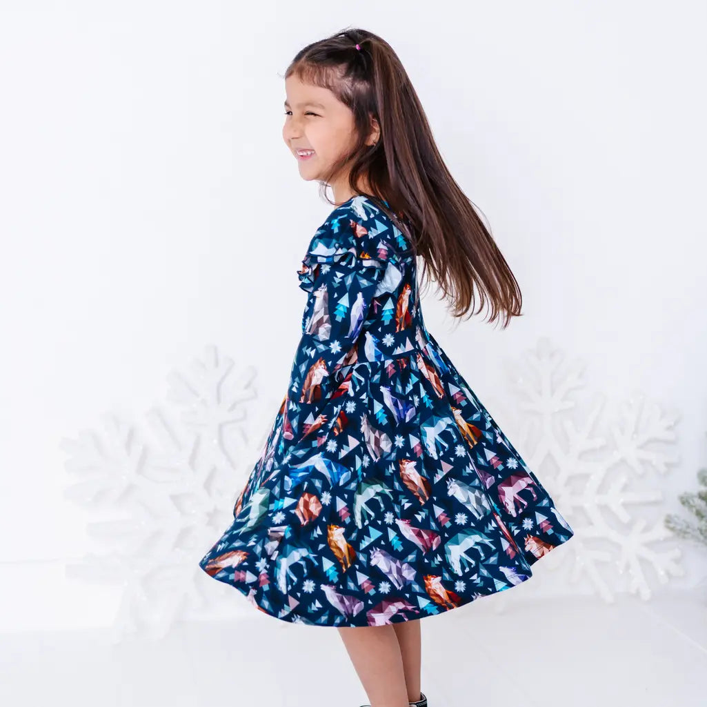 A child twirling sideways and laughing with the full skirt flaring out, in a long sleeve dress with flutter ruffles in a navy print of geometric wolves, triangle pine trees, and snowflakes.