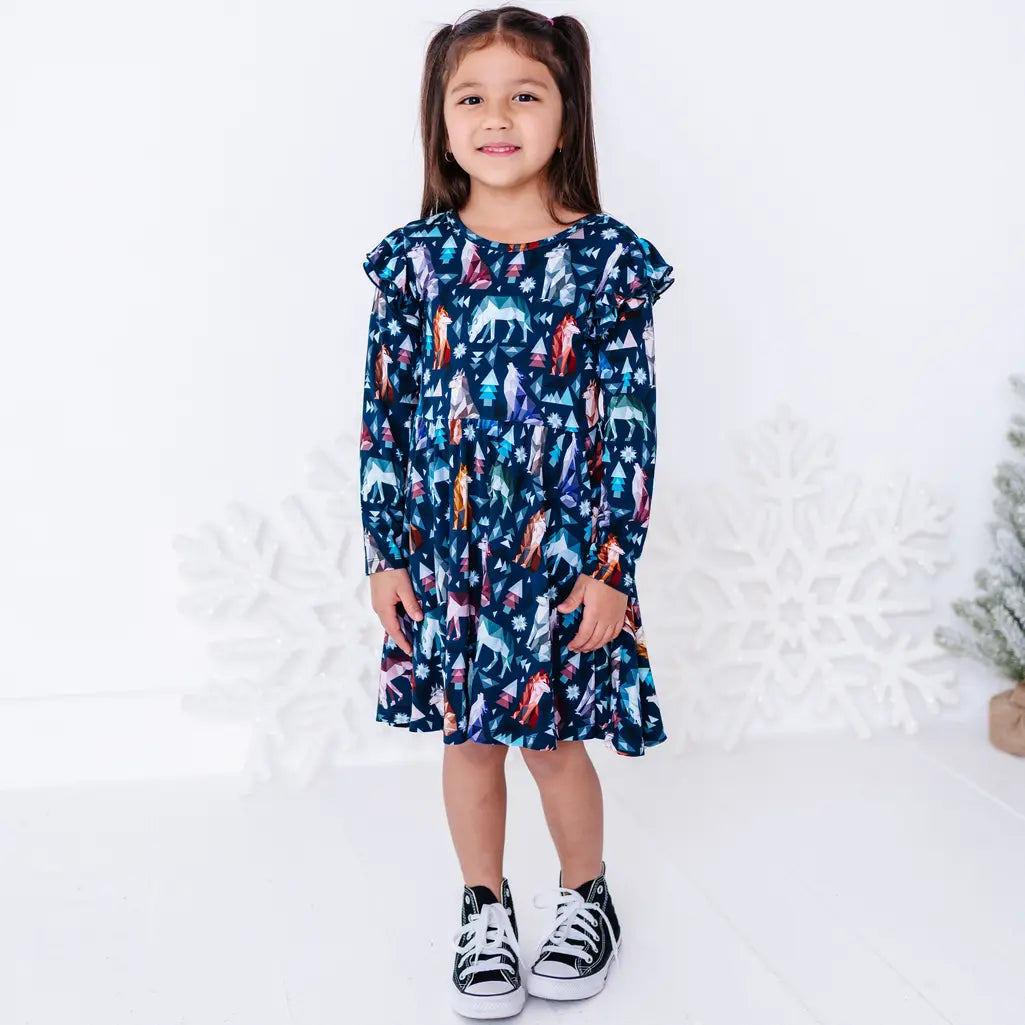 A child smiling and standing with arms at the sides in a long sleeve twirl dress with flutter ruffles in a navy print of geometric wolves, triangle pine trees, and snowflakes.