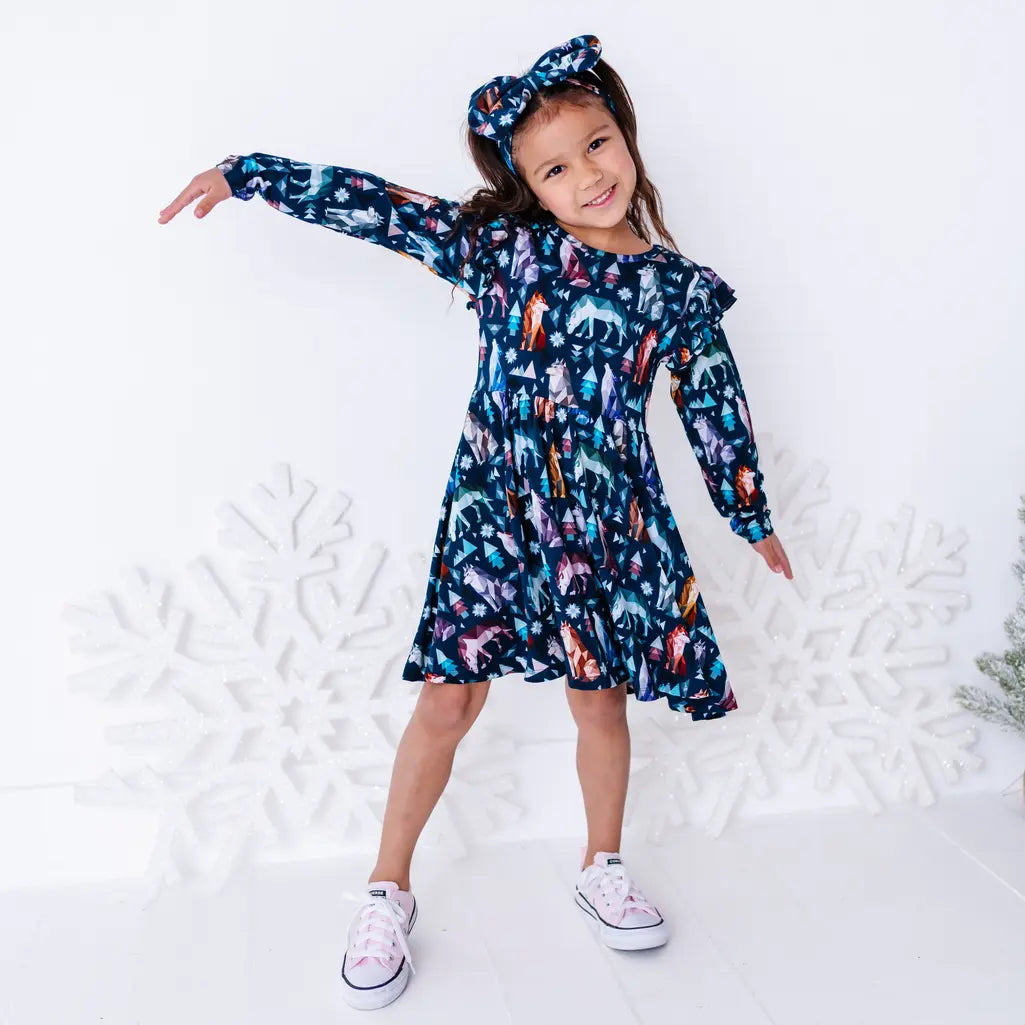 A child dancing with arms stretched out wide and a bow headband, in a long sleeve twirl dress in a navy print of geometric wolves, triangle pine trees, and snowflakes.