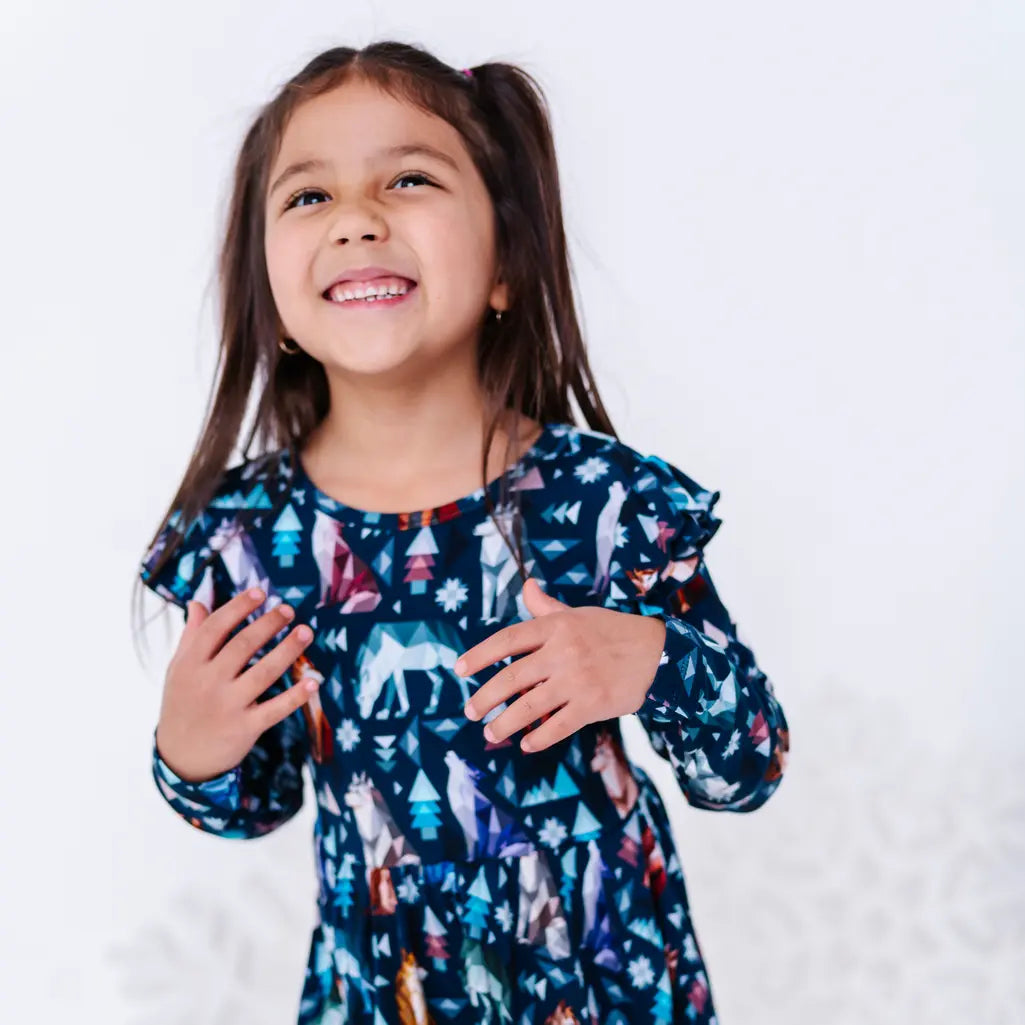 Close portrait of a child smiling and looking up with hands on the chest, in a long sleeve twirl dress with flutter ruffles in a navy print of geometric wolves, triangle pine trees, and snowflakes.