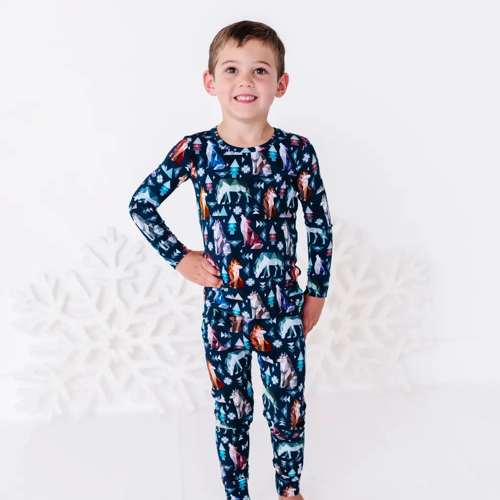 A child smiling with one hand on the hip, standing in a long sleeve pajama top and matching pants in a navy print of geometric wolves, triangle pine trees, and snowflakes.