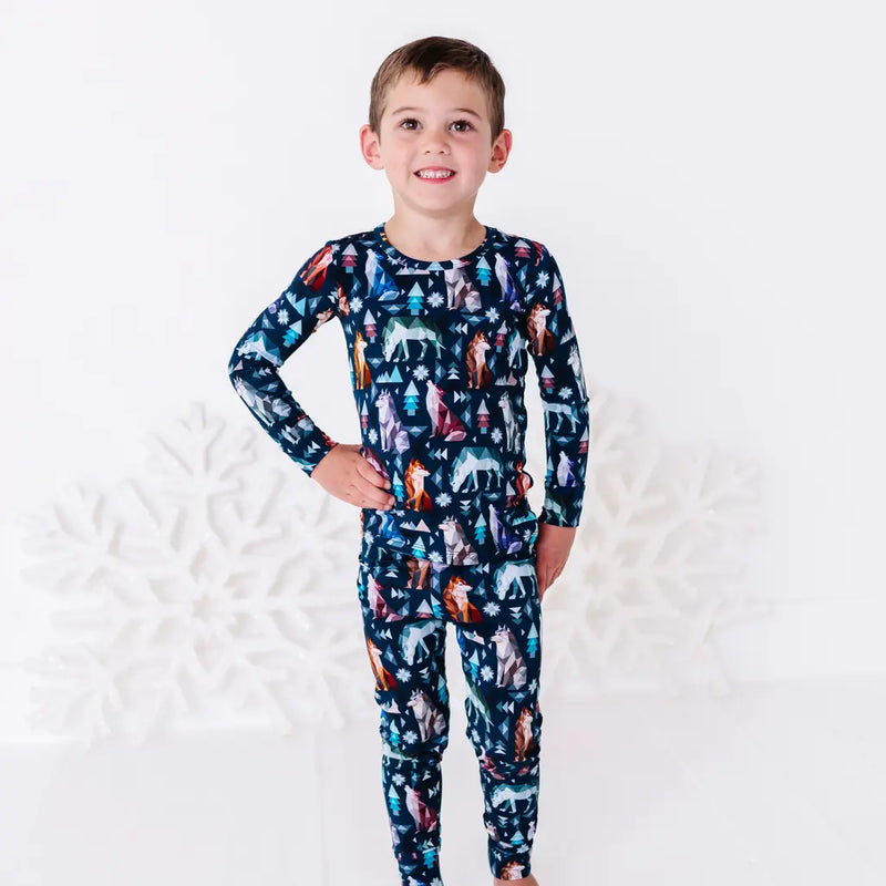 A child smiling with one hand on the hip, standing in a long sleeve pajama top and matching pants in a navy print of geometric wolves, triangle pine trees, and snowflakes.