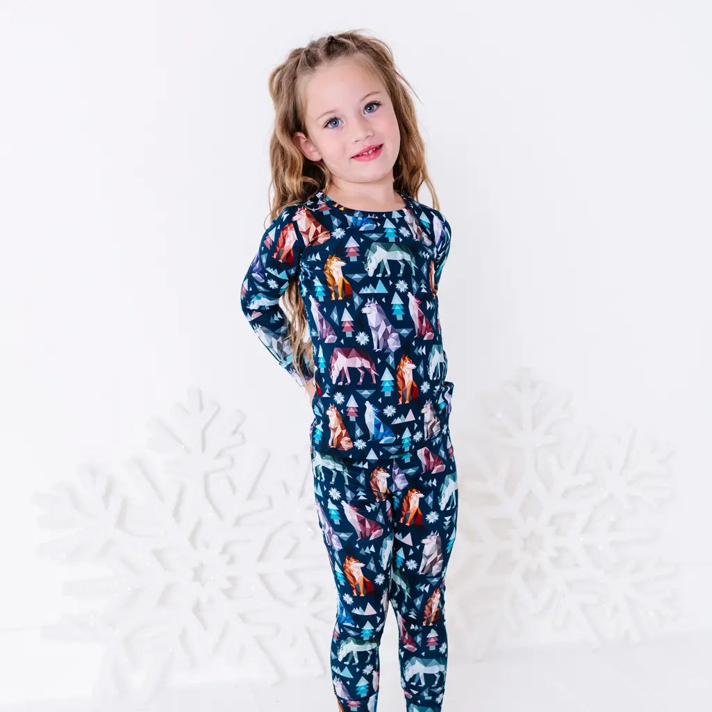 A child turned to the side with a hand on the hip and a soft smile, in a long sleeve pajama top and matching pants in a navy print of geometric wolves, triangle pine trees, and snowflakes.