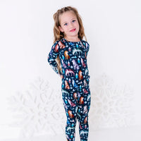 A child turned to the side with a hand on the hip and a soft smile, in a long sleeve pajama top and matching pants in a navy print of geometric wolves, triangle pine trees, and snowflakes.