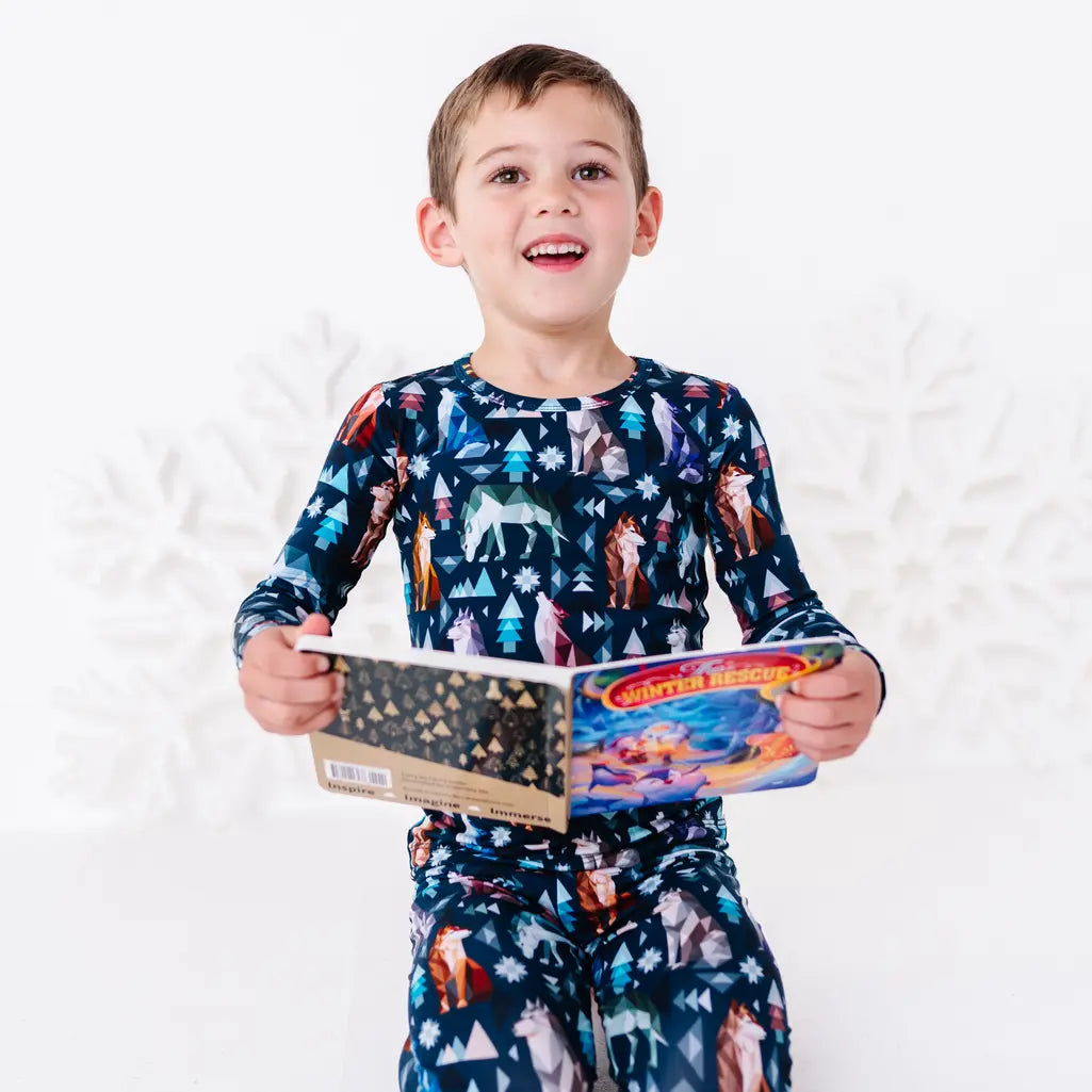 A child smiling at the camera and holding an open board book at the waist, in a long sleeve pajama top in a navy print of geometric wolves, triangle pine trees, and snowflakes.