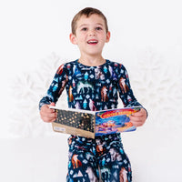 A child smiling at the camera and holding an open board book at the waist, in a long sleeve pajama top in a navy print of geometric wolves, triangle pine trees, and snowflakes.