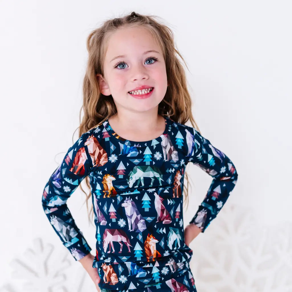 A child with a big smile and hands on the hips, shown from the chest up, in a long sleeve pajama top in a navy print of geometric wolves, triangle pine trees, and snowflakes.