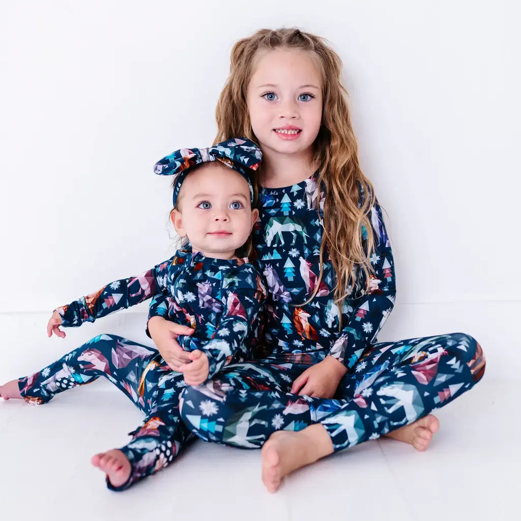 An older sibling sitting with a baby, both looking at the camera, in a pajama set and zip up baby pajama in a navy print of geometric wolves, triangle pine trees, and snowflakes.