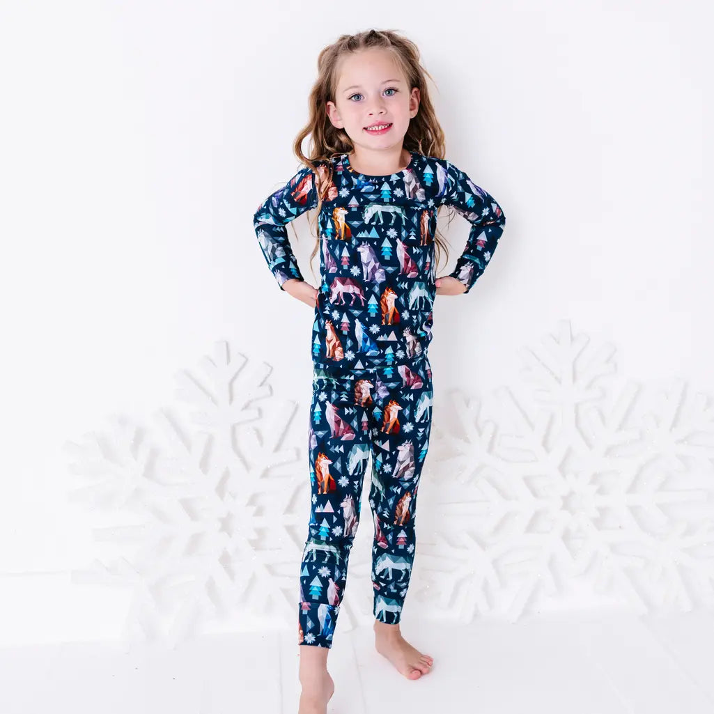 A child standing barefoot with hands on the hips and a small smile, in a long sleeve pajama top and matching pants in a navy print of geometric wolves, triangle pine trees, and snowflakes.