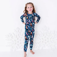 A child standing barefoot with hands on the hips and a small smile, in a long sleeve pajama top and matching pants in a navy print of geometric wolves, triangle pine trees, and snowflakes.