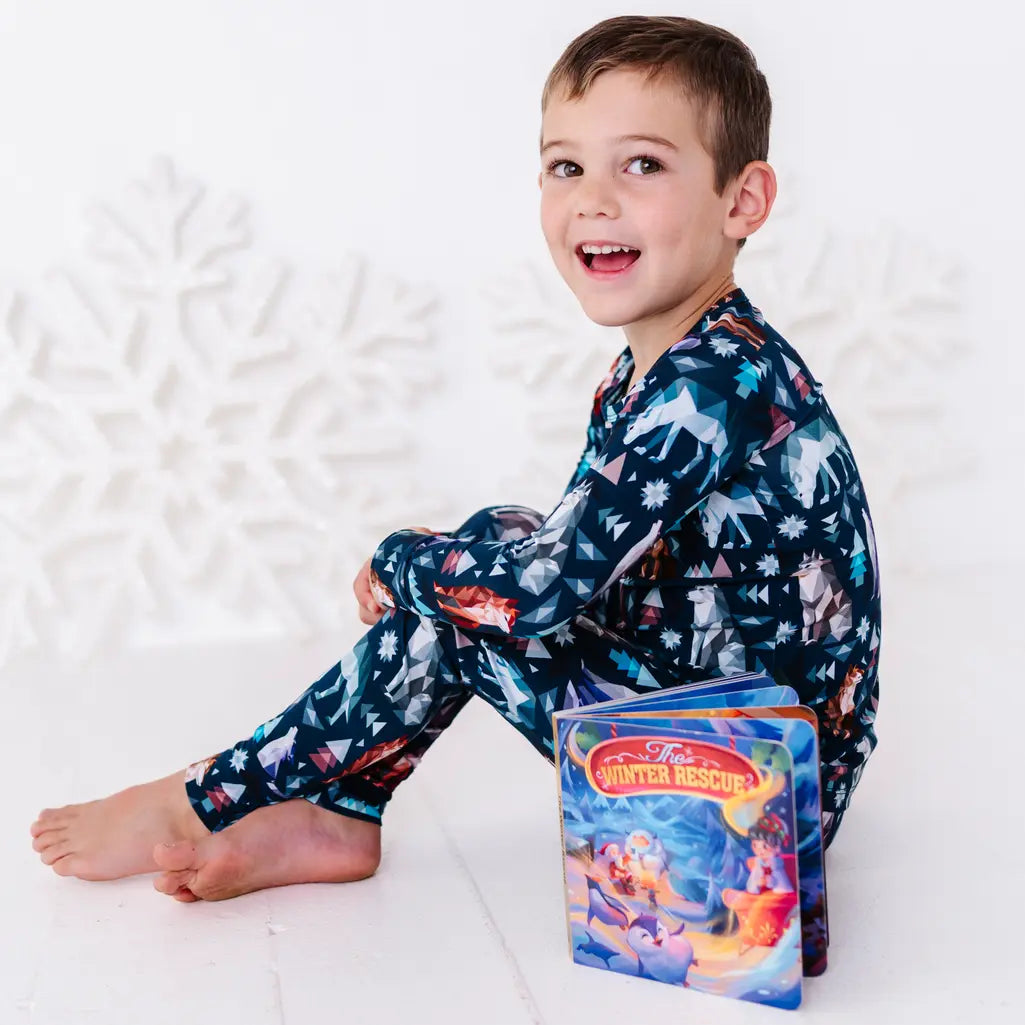 A child sitting barefoot and smiling at the camera with a board book beside them, in a long sleeve pajama set in a navy print of geometric wolves, triangle pine trees, and snowflakes.