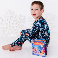 A child sitting barefoot and smiling at the camera with a board book beside them, in a long sleeve pajama set in a navy print of geometric wolves, triangle pine trees, and snowflakes.