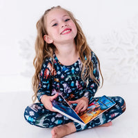 A child sitting cross legged with an open board book on the lap and a big smile, in a long sleeve pajama set in a navy print of geometric wolves, triangle pine trees, and snowflakes.
