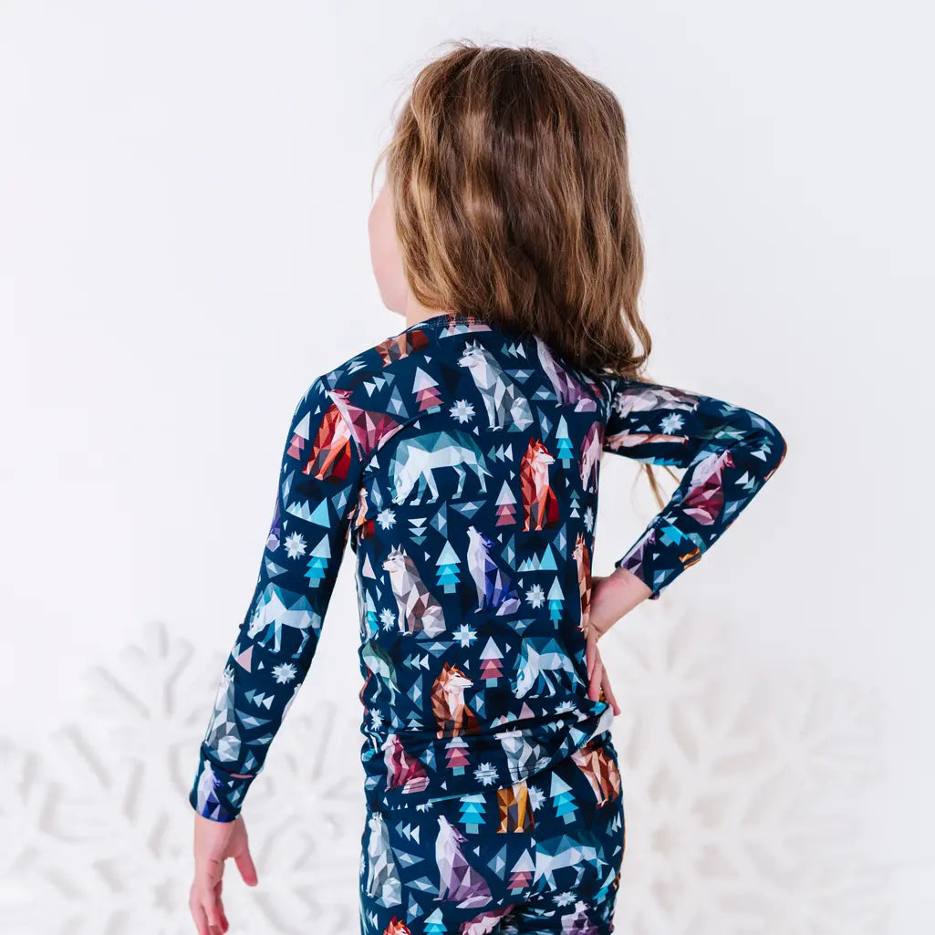 Back view of a child with a hand on the hip in a long sleeve pajama top and matching pants in a navy print of geometric wolves, triangle pine trees, and snowflakes.