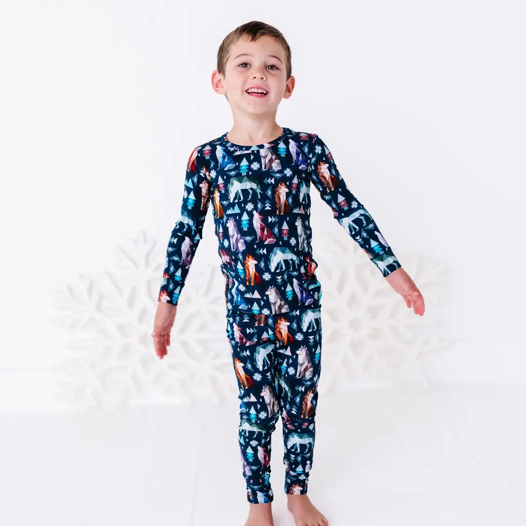 A child standing with arms stretched out and a big smile, shown to the knees, in a long sleeve pajama top and matching pants in a navy print of geometric wolves, triangle pine trees, and snowflakes.