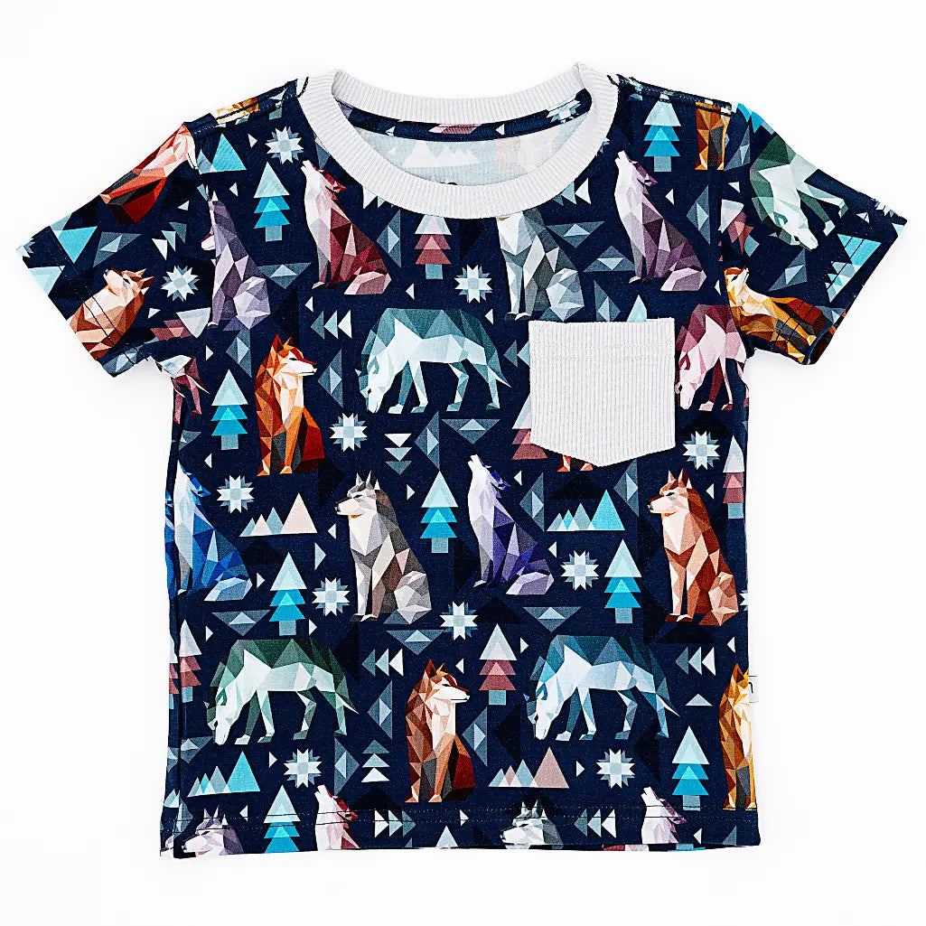 Flat lay of a short sleeve pocket tee with a cream ribbed collar and pocket in a navy print of geometric wolves, triangle pine trees, and snowflakes on a white background.