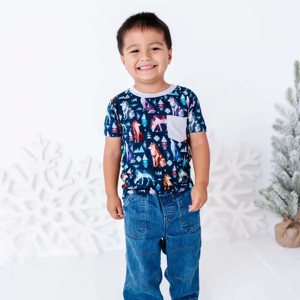A child standing and smiling in jeans and a short sleeve pocket tee with a cream ribbed collar and pocket in a navy print of geometric wolves, triangle pine trees, and snowflakes.