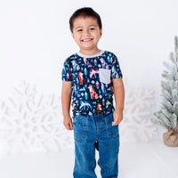 A child standing and smiling in jeans and a short sleeve pocket tee with a cream ribbed collar and pocket in a navy print of geometric wolves, triangle pine trees, and snowflakes.
