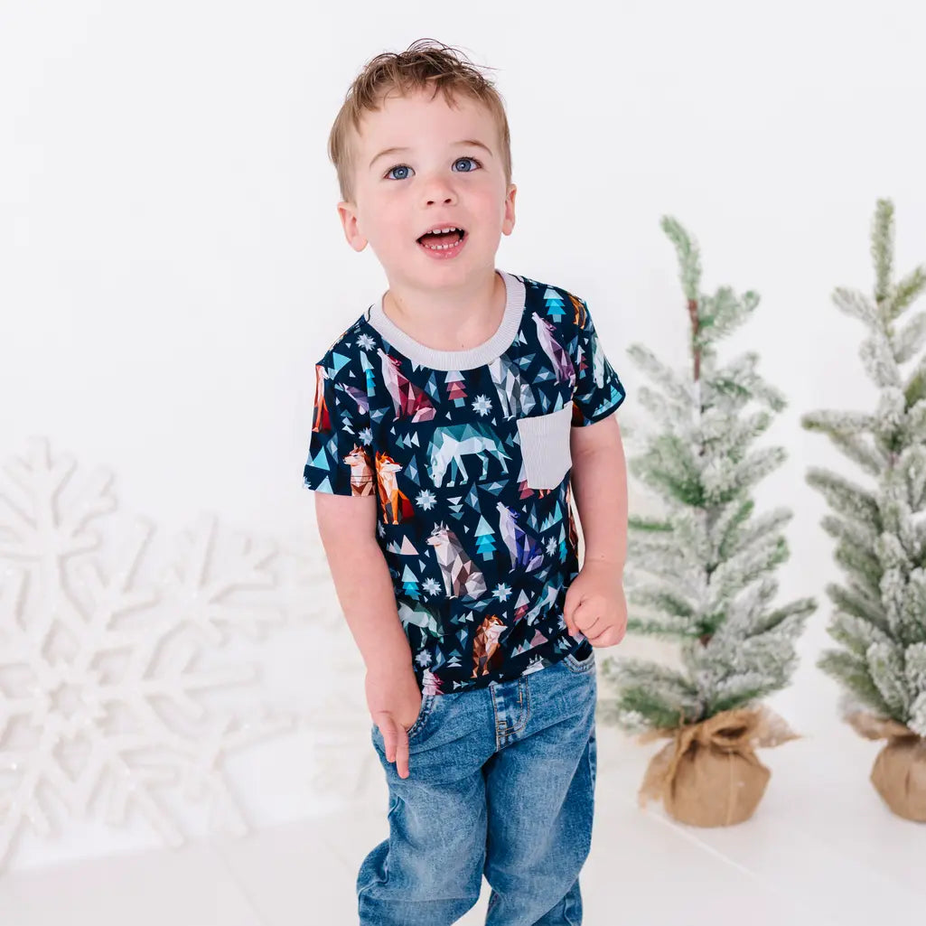 A child with hands in the jeans pockets and an open mouth smile, in a short sleeve pocket tee in a navy print of geometric wolves, triangle pine trees, and snowflakes.
