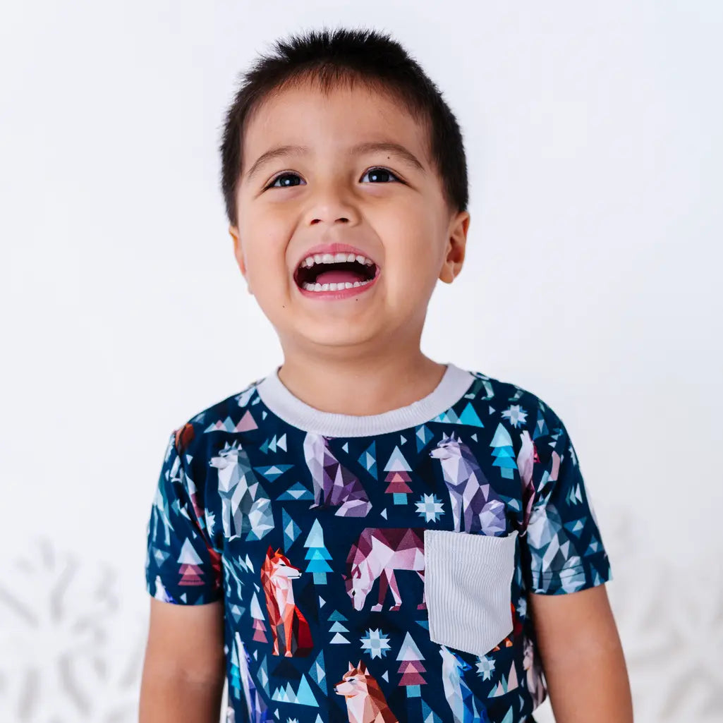 A child laughing with an open mouth in a short sleeve pocket tee with a cream ribbed collar and pocket in a navy print of geometric wolves, triangle pine trees, and snowflakes.