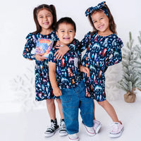 Three siblings leaning in and smiling at the camera in two long sleeve twirl dresses and a pocket tee in a navy print of geometric wolves, triangle pine trees, and snowflakes.