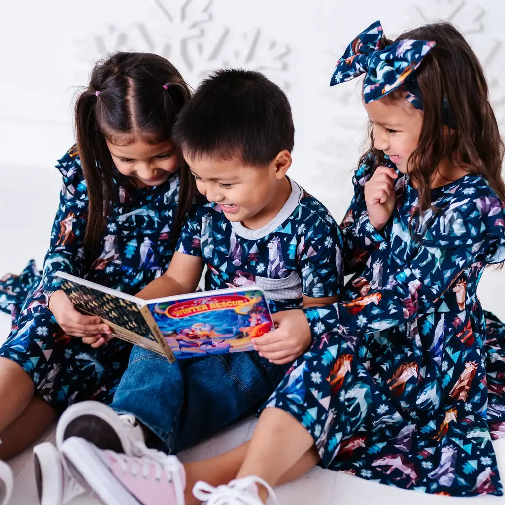 Three siblings sitting close and reading a storybook, in two long sleeve twirl dresses and a pocket tee in a navy print of geometric wolves, triangle pine trees, and snowflakes.