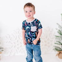 A child standing in jeans with a soft smile and one hand at the hip, in a short sleeve pocket tee in a navy print of geometric wolves, triangle pine trees, and snowflakes.