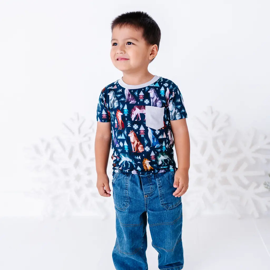 A child standing in jeans and looking off to the side, in a short sleeve pocket tee with a cream ribbed collar and pocket in a navy print of geometric wolves, triangle pine trees, and snowflakes.