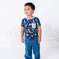 A child standing in jeans and looking off to the side, in a short sleeve pocket tee with a cream ribbed collar and pocket in a navy print of geometric wolves, triangle pine trees, and snowflakes.