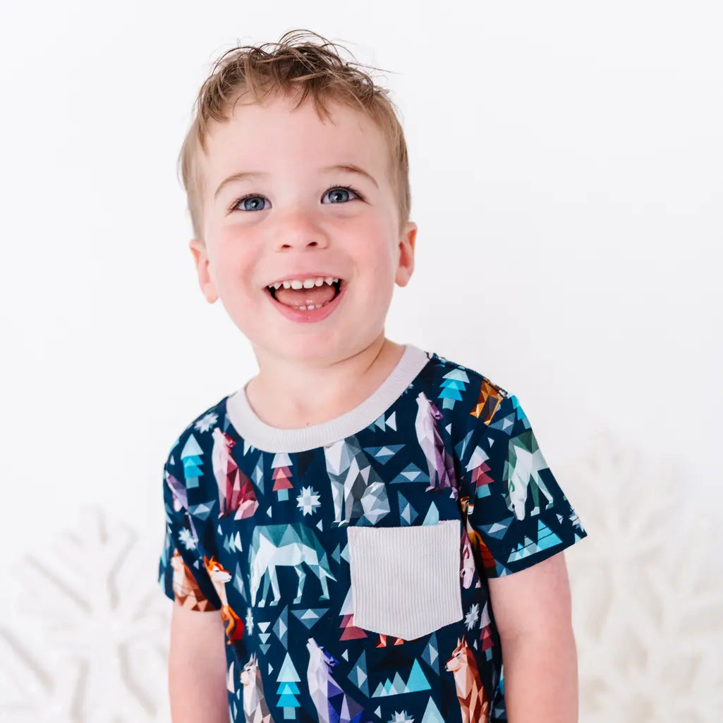 A child laughing with the head tilted, shown from the chest up, in a short sleeve pocket tee in a navy print of geometric wolves, triangle pine trees, and snowflakes.