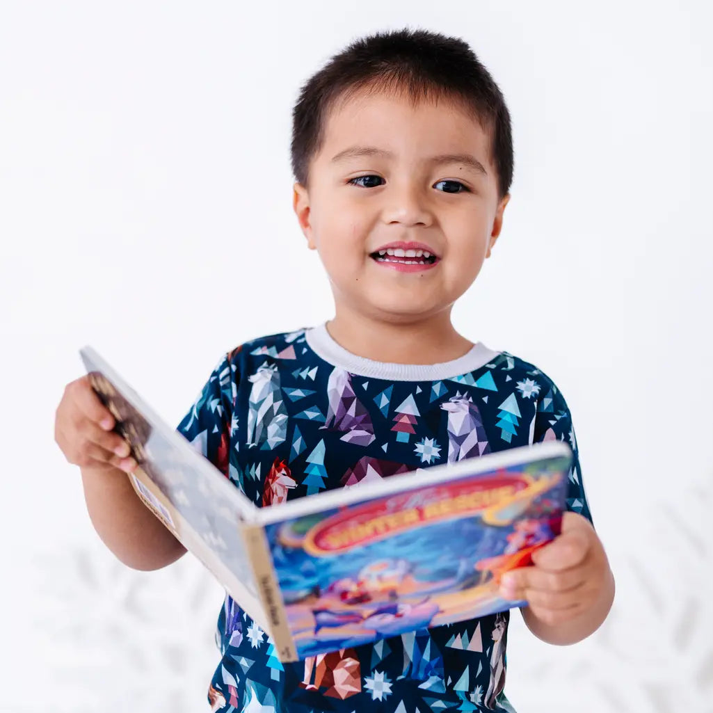 A child smiling and holding an open board book, shown from the chest up, in a short sleeve pocket tee in a navy print of geometric wolves, triangle pine trees, and snowflakes.