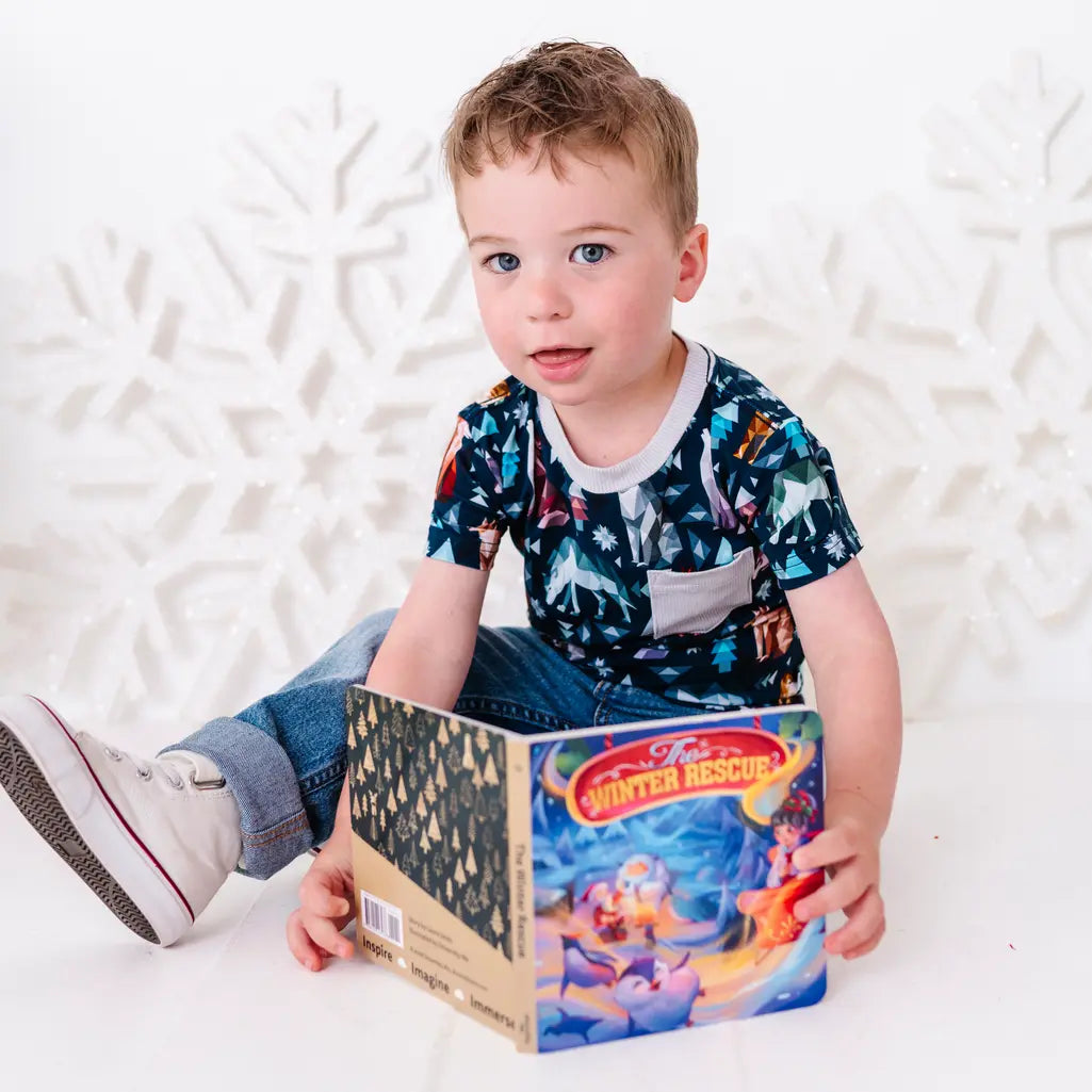 A child sitting on a white floor with a board book, looking at the camera, in jeans and a short sleeve pocket tee in a navy print of geometric wolves, triangle pine trees, and snowflakes.