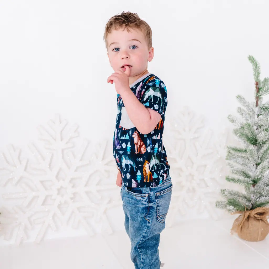 Side view of a child standing in jeans with a finger at the mouth, in a short sleeve pocket tee in a navy print of geometric wolves, triangle pine trees, and snowflakes.