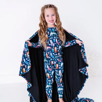 A child smiling at the camera and holding out the sides of a navy blanket with a black reverse in a geometric wolf print with triangle pine trees and snowflakes.