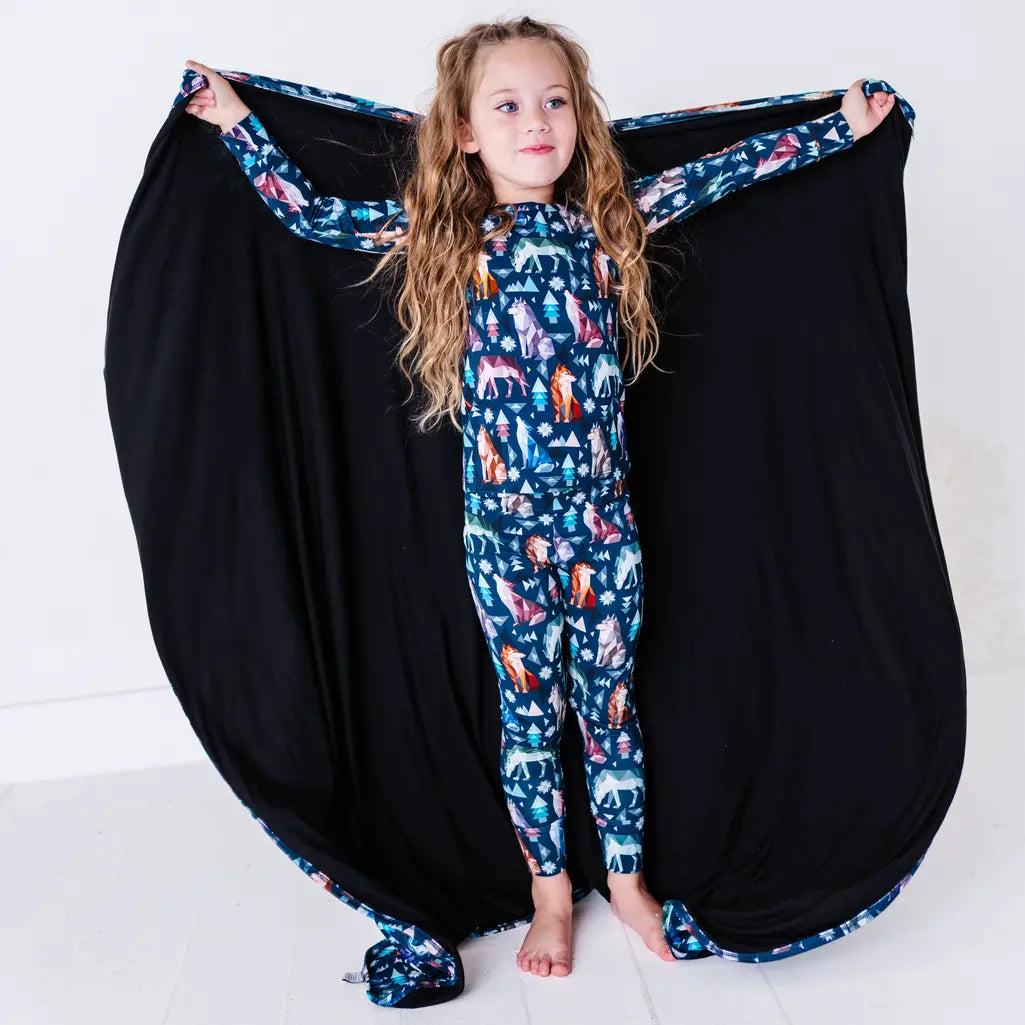 A child standing calmly with the arms stretched wide, holding out a navy blanket with a black reverse in a geometric wolf print with triangle pine trees and snowflakes like wings.