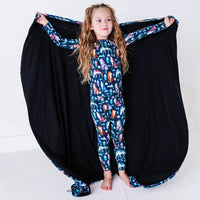 A child standing calmly with the arms stretched wide, holding out a navy blanket with a black reverse in a geometric wolf print with triangle pine trees and snowflakes like wings.