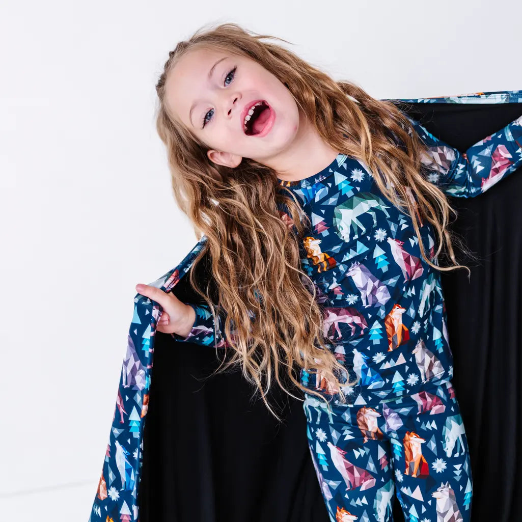 A child laughing with the head tilted and one hand holding the edge of a navy blanket with a black reverse in a geometric wolf print with triangle pine trees and snowflakes.