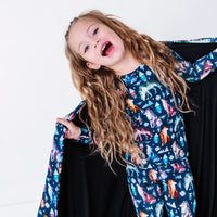 A child laughing with the head tilted and one hand holding the edge of a navy blanket with a black reverse in a geometric wolf print with triangle pine trees and snowflakes.