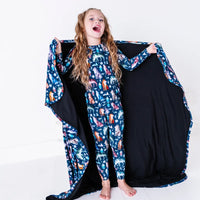 A laughing child holding out a navy blanket with a black reverse in a geometric wolf print with triangle pine trees and snowflakes like a cape.