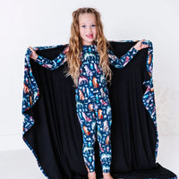 A smiling child with the arms out at shoulder height and a navy blanket with a black reverse in a geometric wolf print with triangle pine trees and snowflakes spread behind.