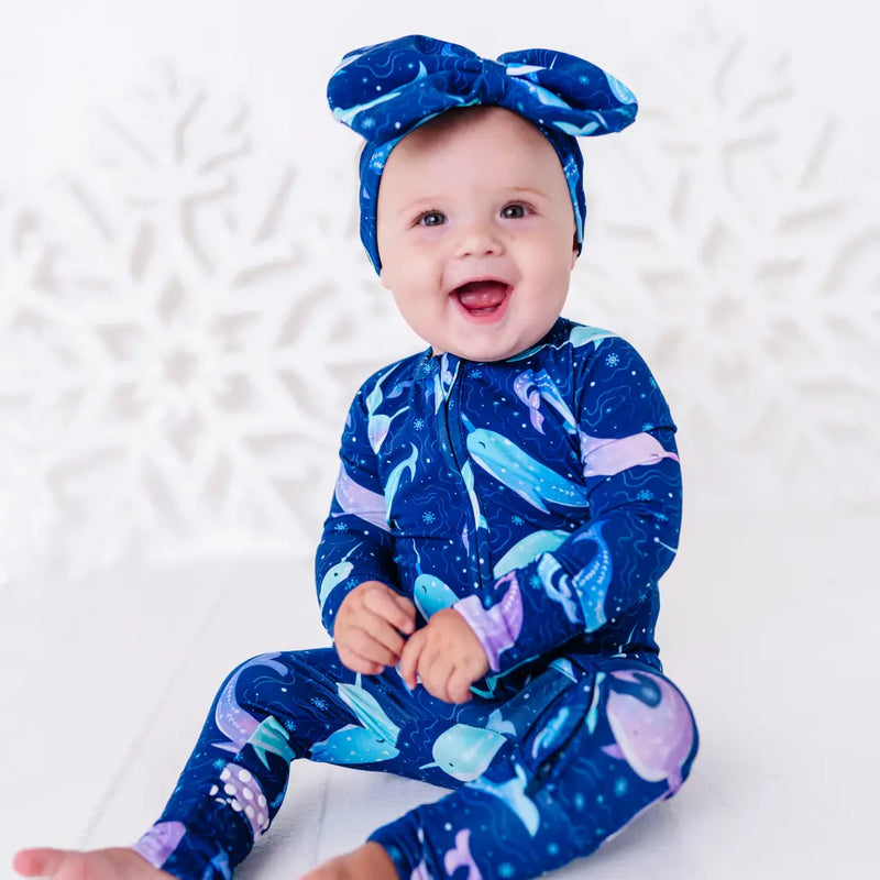 A baby with a big open mouth smile sitting on a white floor in a zip up pajama and bow headband in a royal blue print of blue and lavender narwhals.