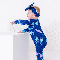 A baby in a bow headband laughing in profile while standing at a white block with one arm stretched out, in a zip up pajama in a royal blue print of blue and lavender narwhals.