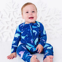 A smiling baby sitting barefoot on a white floor and looking toward the camera in a zip up pajama in a royal blue print of blue and lavender narwhals.