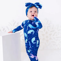 A smiling baby in a bow headband standing at a white block with a hand to the mouth, in a zip up pajama in a royal blue print of blue and lavender narwhals.