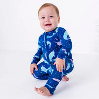 A smiling baby crouching on a white floor with one bare foot stretched out, in a zip up pajama in a royal blue print of blue and lavender narwhals.