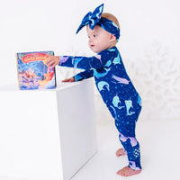 A baby in a bow headband standing and reaching toward a storybook on a white block, in a zip up pajama in a royal blue print of blue and lavender narwhals.